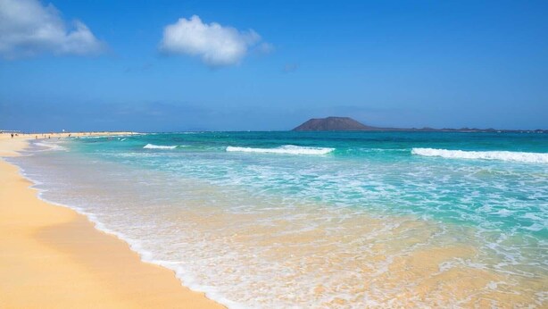 A beach with clear turquoise water, a sandy shore, and a mountain in the distance.