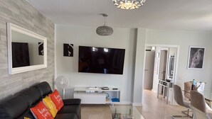 A modern living room with a black sofa, a glass coffee table, a flat-screen TV, and a dining area with chairs and a table.