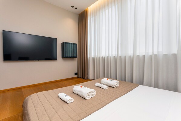 Habitación de hotel con cama, televisor de pantalla plana, televisor montado en la pared y ventana con cortinas.