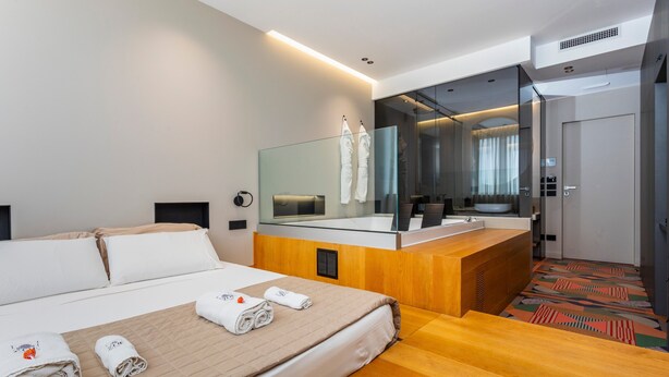 Ein modernes Hotelzimmer mit Bett, Schreibtisch aus Holz, großem Spiegel und abgetrenntem Badezimmer.