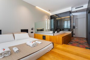 Una habitación de hotel moderna con una cama, un escritorio de madera, un espejo grande y un baño visible a través de una mampara de vidrio.