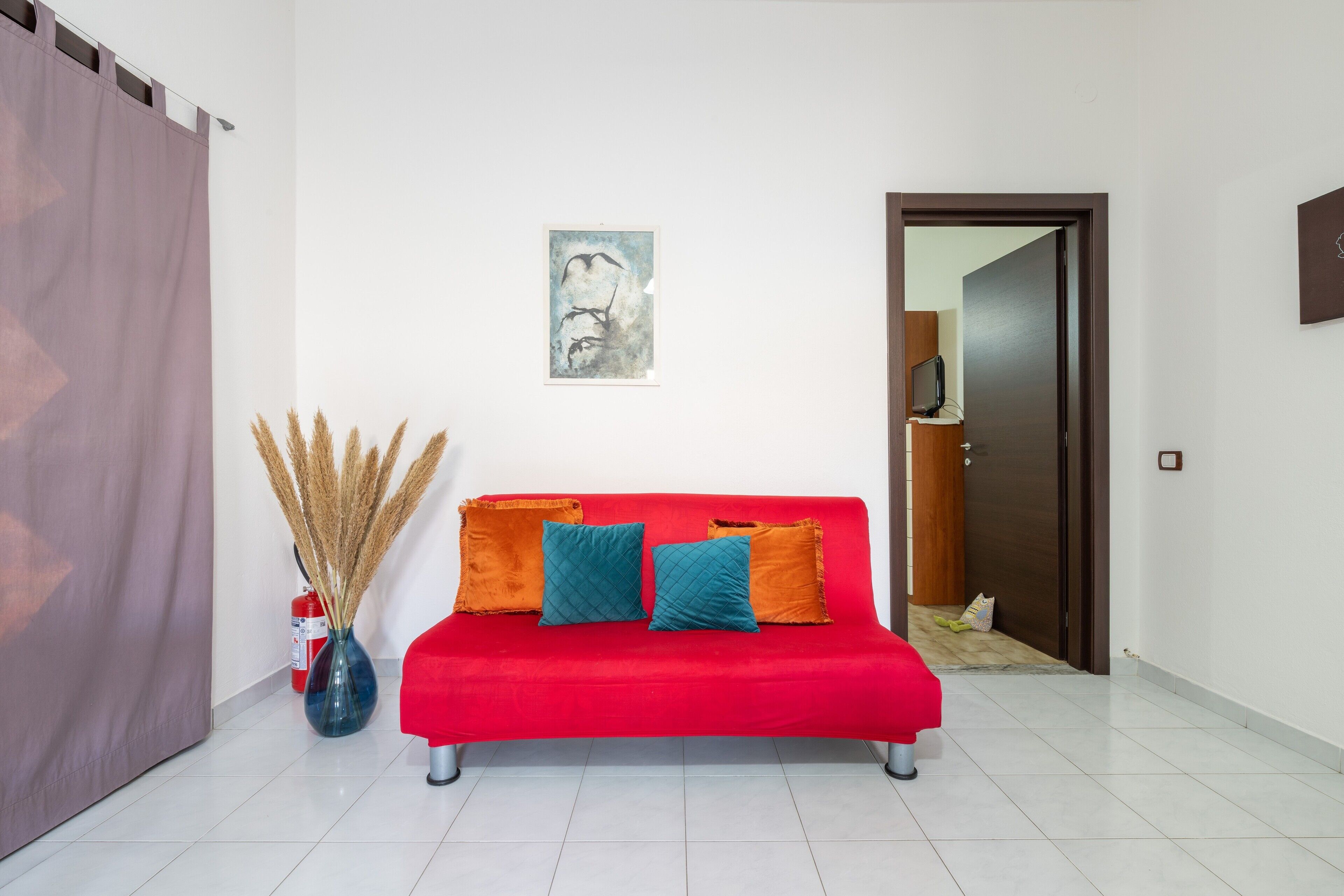 Un sofá rojo con tres cojines, un jarrón con hierbas altas y una obra enmarcada en la pared.