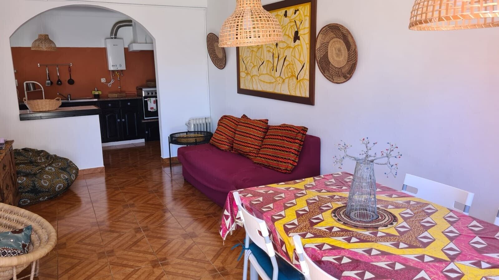 A cozy living area with a sofa, a dining table with a patterned tablecloth, and a kitchen visible in the background.