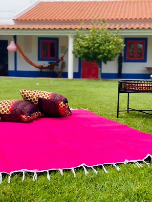 En græsplæne med et rødt tæppe, mønstrede puder, en hængekøje og en bygning med et tag af fliser og blå og hvide vægge.
