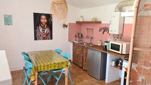 Et lille køkken med vask, mikroovn og køleskab. Spisebord med stole og en hængende lampe.