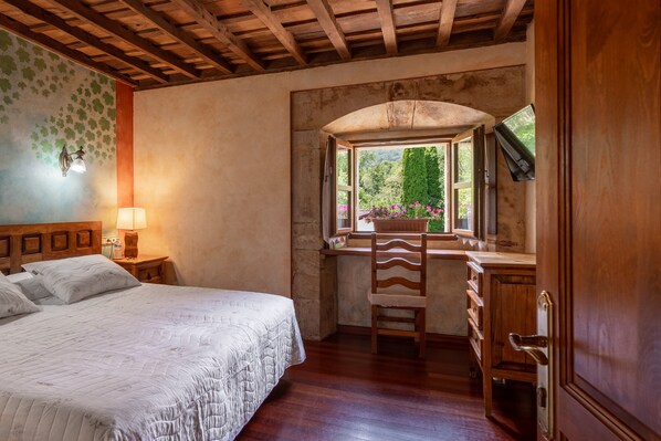 A bedroom with a bed, a desk, a chair, a window with a view of greenery, and a wooden ceiling.