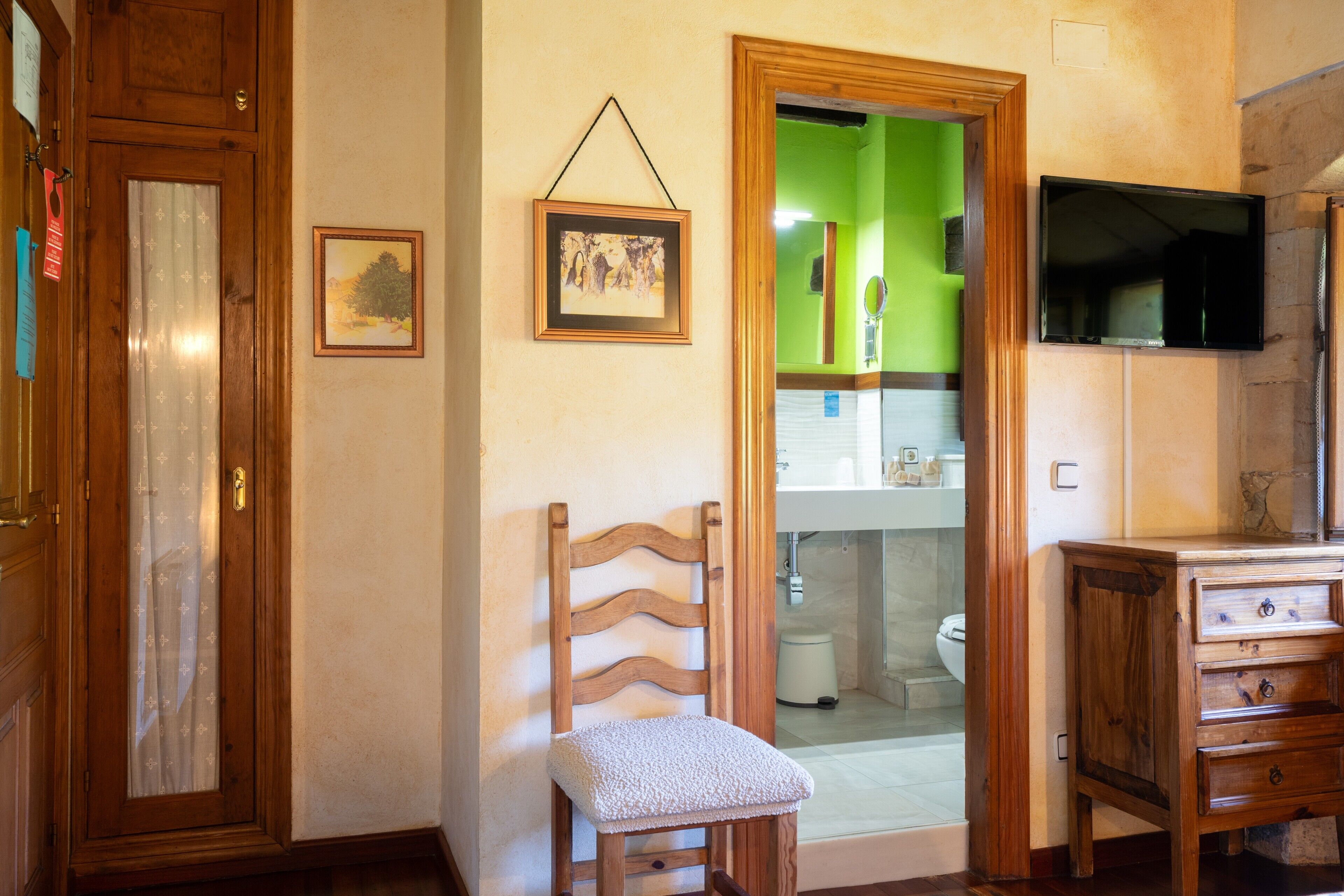 Una habitación con una silla de madera, un armario de madera, un cuadro enmarcado en la pared y una puerta verde que da a un baño.