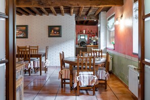 A dining area with wooden tables and chairs, a brick wall with framed artwork, and a kitchen area with a red wall and white cabinets. - Room 'Araucaria' with Mountain View, Shared Garden and Wi-Fi (Con)