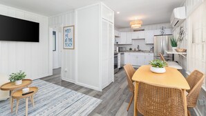 Une cuisine moderne avec des meubles blancs, un coin repas avec des chaises en osier et un téléviseur à écran plat fixé au mur.