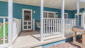 Un porche couvert avec des balustrades blanches, une porte ornée d’une pancarte « BIENVENUE » et un espace détente avec des chaises et une table.