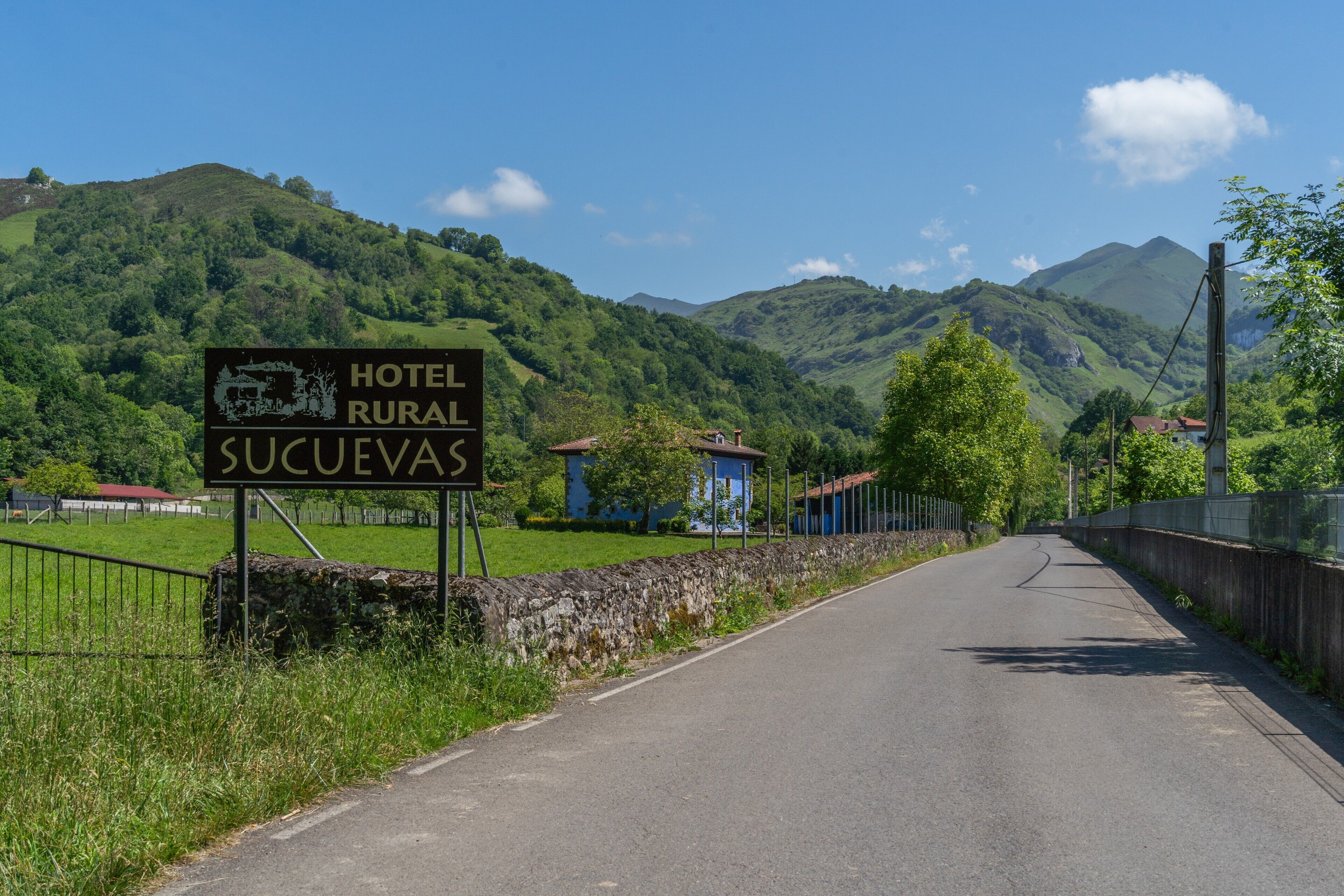 A rural road leading to a hotel named 'Hotel Rural Sucuevas' with a stone wall and greenery on one side.