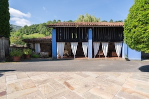 Un bâtiment avec un toit recouvert de tuiles, une véranda ombragée avec des rideaux blancs, des murs en pierre et des plantes en pot.