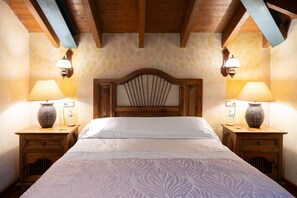 A bedroom with a wooden headboard, two bedside tables with lamps, a patterned bedspread, and a wall with a leaf pattern.