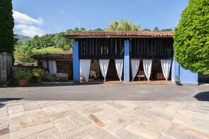 A building with a tiled roof, a covered porch with white curtains, stone walls, and potted plants.