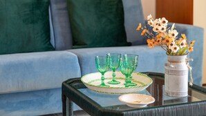 Un salón con un sofá azul, una mesa de centro de vidrio con una bandeja que contiene vasos verdes, un jarrón blanco con flores y un plato blanco pequeño.
