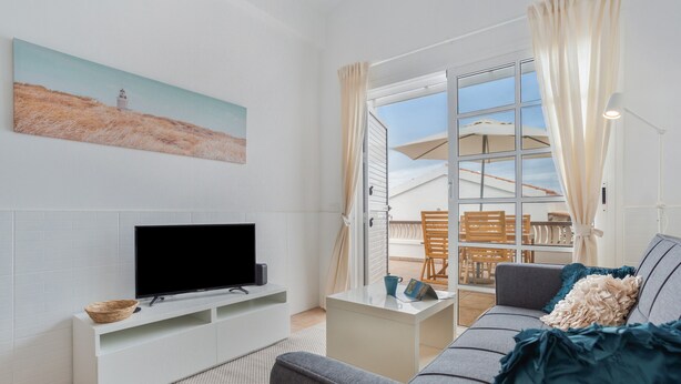 A living room with a flat-screen TV, a white entertainment unit, a grey sofa, a small coffee table, and a large window with curtains.