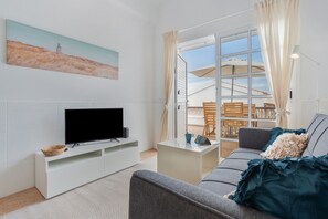 Una sala de estar con televisor de pantalla plana, mueble blanco para equipo audiovisual, sofá gris, una mesita de centro pequeña y un ventanal con cortinas.