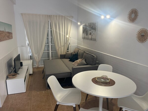 Un espacio habitable compacto con sofá, una mesa de comedor con sillas, televisor y ventana con cortinas.