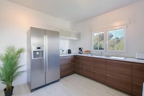A modern kitchen with a stainless steel refrigerator, wooden cabinets, a white countertop, and a window with a view of greenery.
