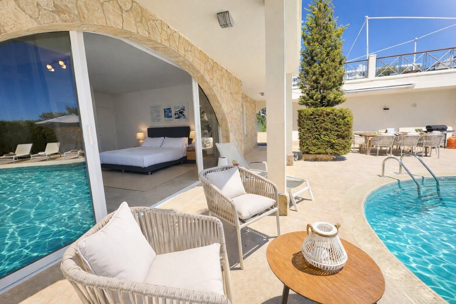 A hotel room with a bed, a balcony with a pool, and outdoor seating.
