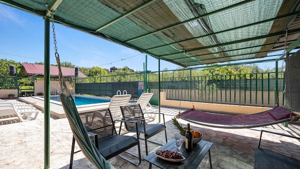 A covered outdoor area with a pool, lounge chairs, and a table with a bowl of fruit and a bottle.