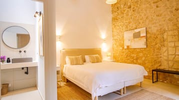 A bedroom with a stone wall, a bed with white bedding, a bedside table with a lamp, and a nightstand.