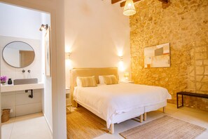 A bedroom with a stone wall, a bed with white bedding, a bedside table with a lamp, and a nightstand.