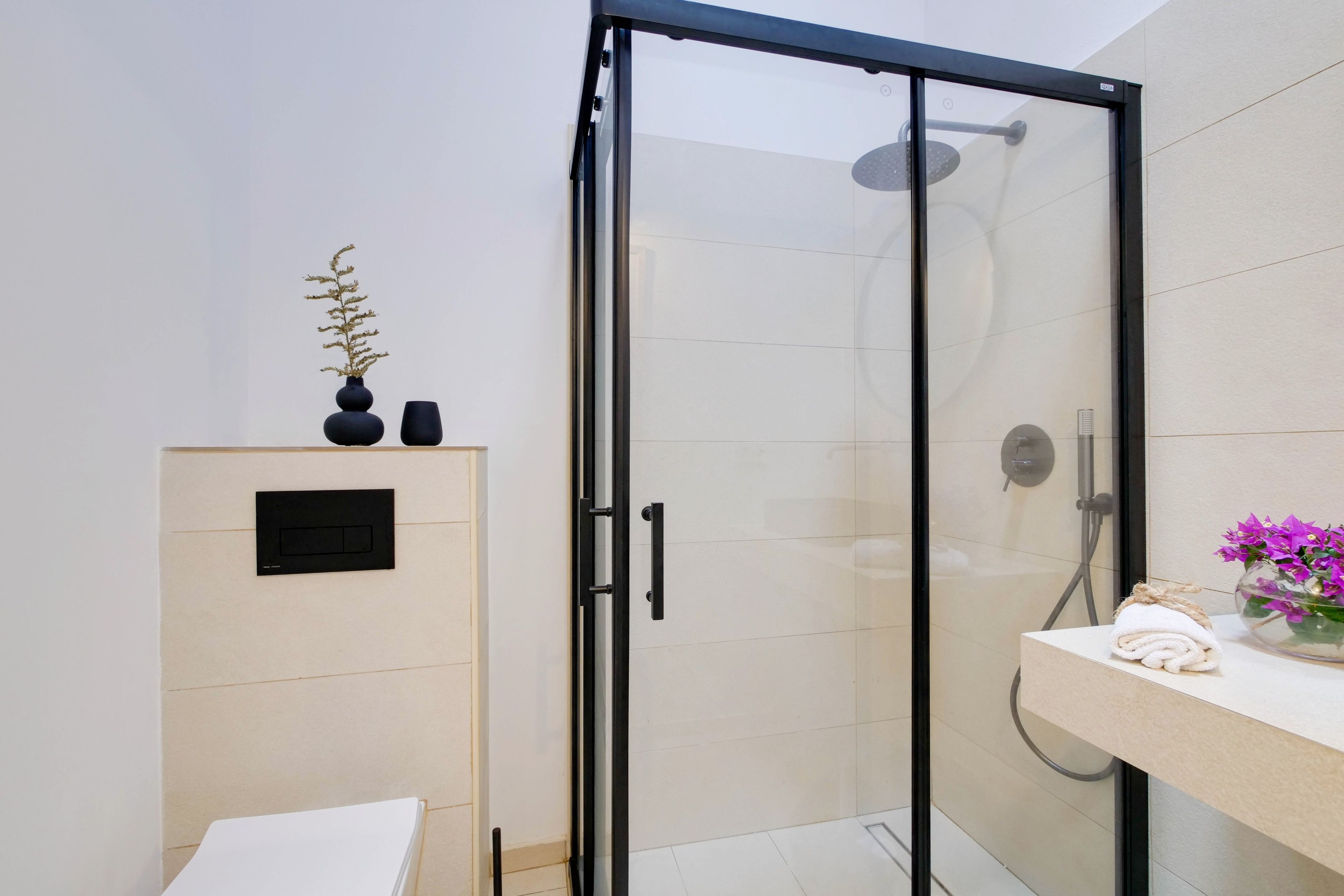 A modern bathroom with a glass shower enclosure, a white sink with a vase of flowers, and a small shelf with decorative items.