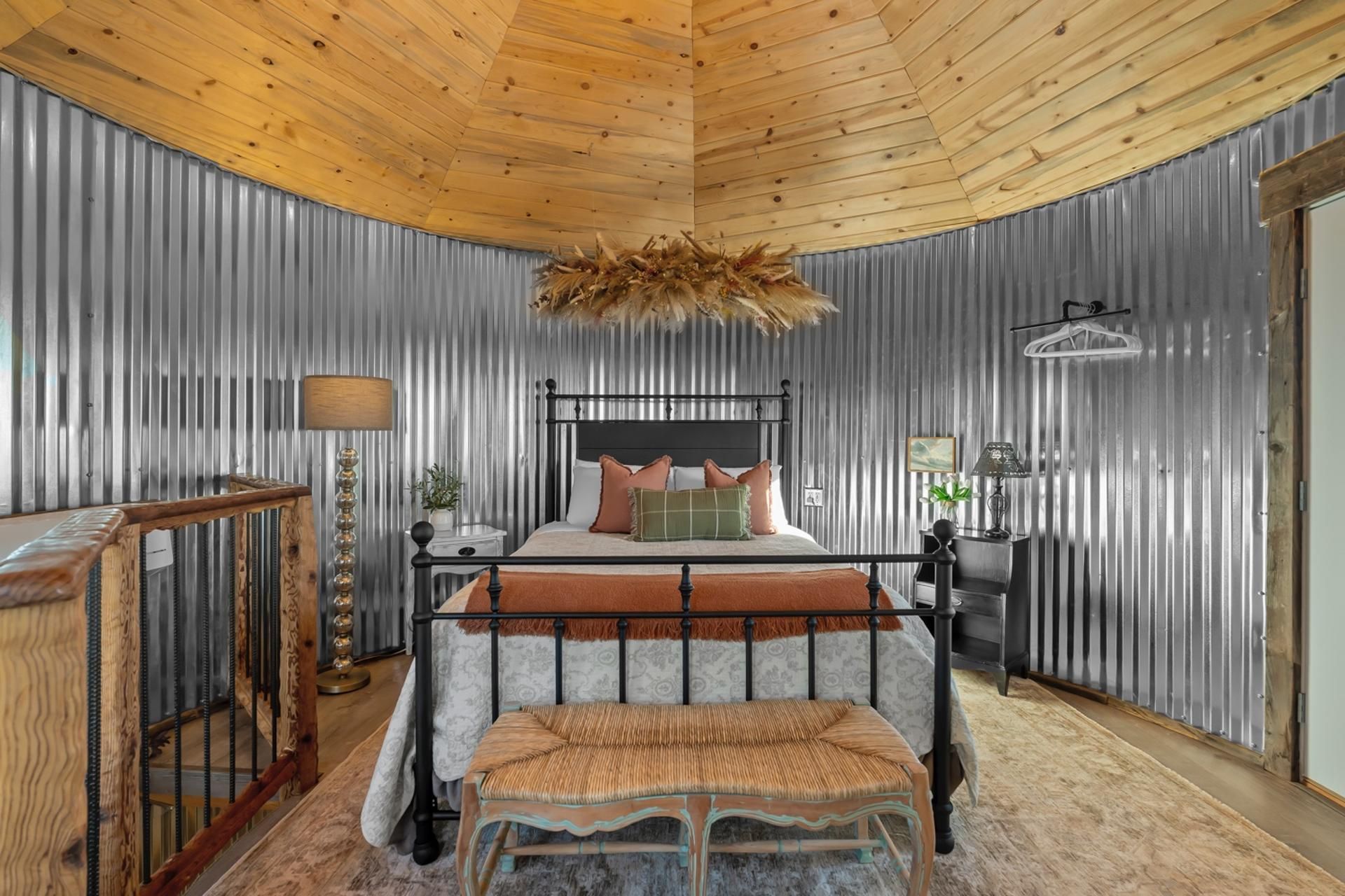 Un dormitorio con techo curvo, una cama con cabecera, un banco, una mesita de noche, una lámpara y un aplique de pared.