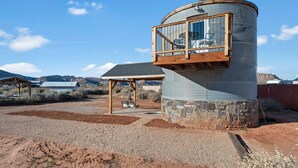 A cylindrical building with a wooden deck and a stone base, surrounded by a gravel area and sparse vegetation. - Southwest Silo Stay- 1 Bedroom Near Zion & Bryce (Fredonia)
