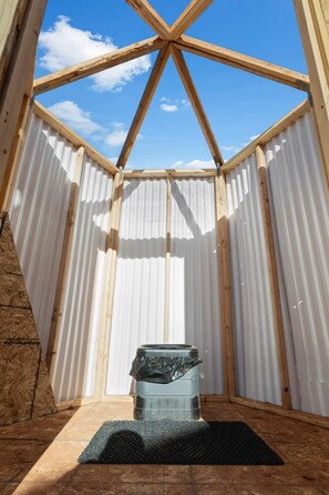 A small, enclosed space with a wooden ceiling and corrugated metal walls, featuring a trash bin and a rug on the floor.