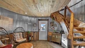 A rustic interior with a wooden ceiling, a metal-framed door, a wooden staircase, and a seating area with a round table and two wicker chairs. - Southwest Silo Stay- 1 Bedroom Near Zion & Bryce (Fredonia)