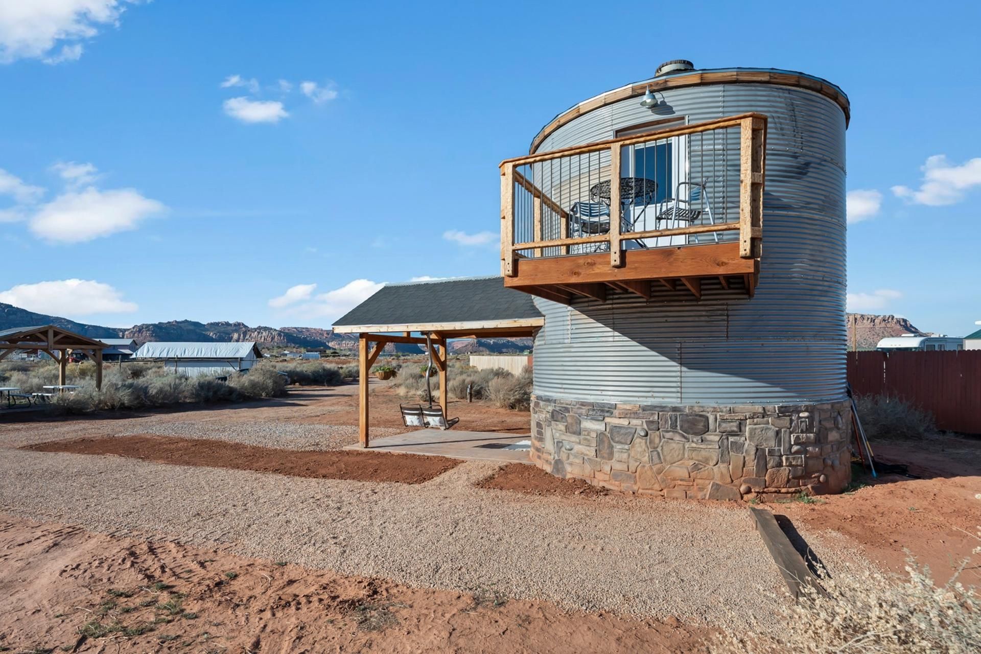 Un bâtiment cylindrique doté d’un balcon en bois, entouré d’une zone recouverte de gravier et d’une végétation clairsemée.