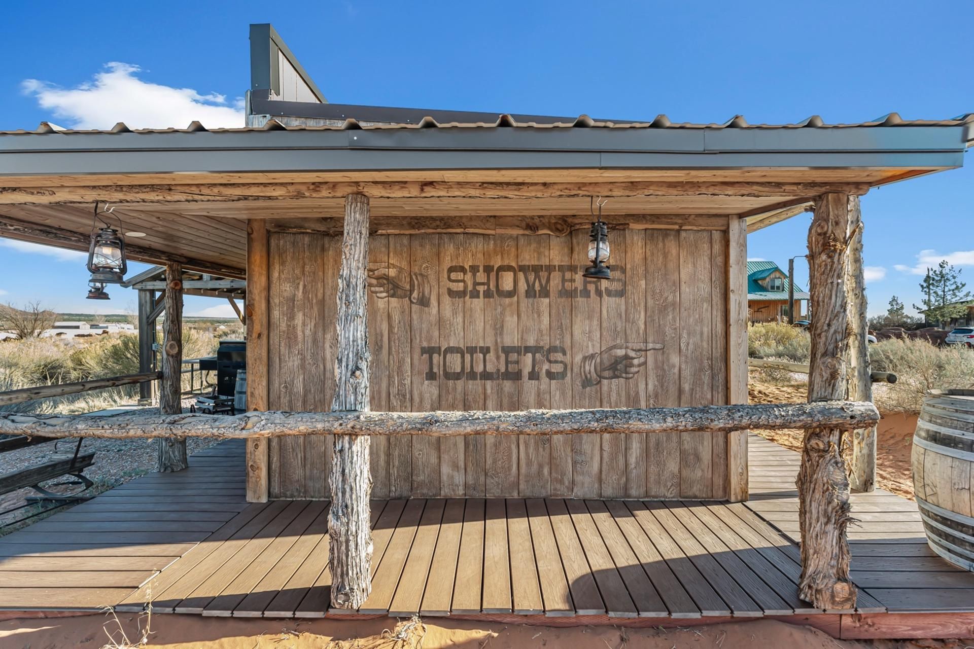 Estructura de madera con la inscripción 'SHOWERS TOILETS' tallada, sostenida por postes de madera, techo metálico y una cubierta de madera al frente.