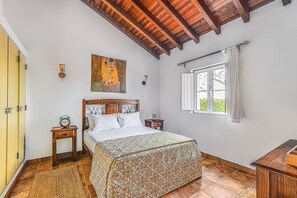 A bedroom with a bed, a nightstand, a wall clock, a painting, a window with curtains, and a wooden ceiling.