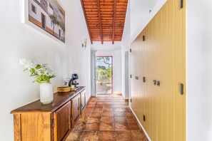 Un pasillo con techo de madera, suelo de baldosas, un mueble de madera con un jarrón de flores y vista a un área exterior a través de una puerta abierta.