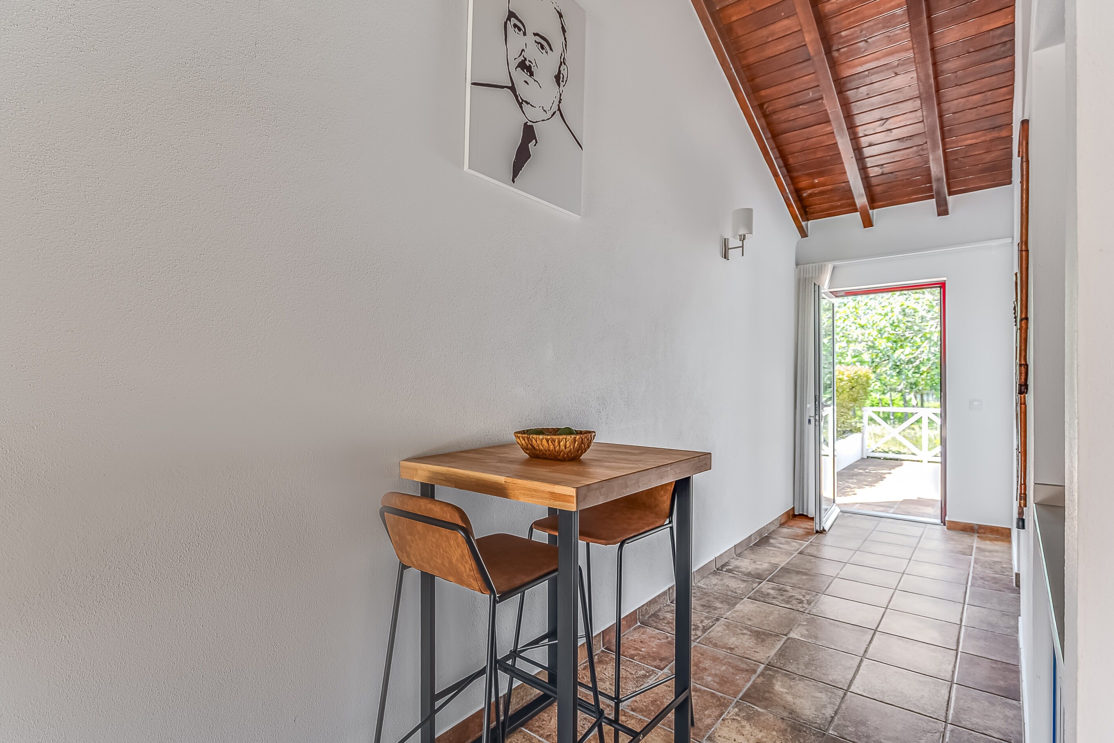 A hallway with a wooden table and two chairs, a framed picture on the wall, and a view of the outside.