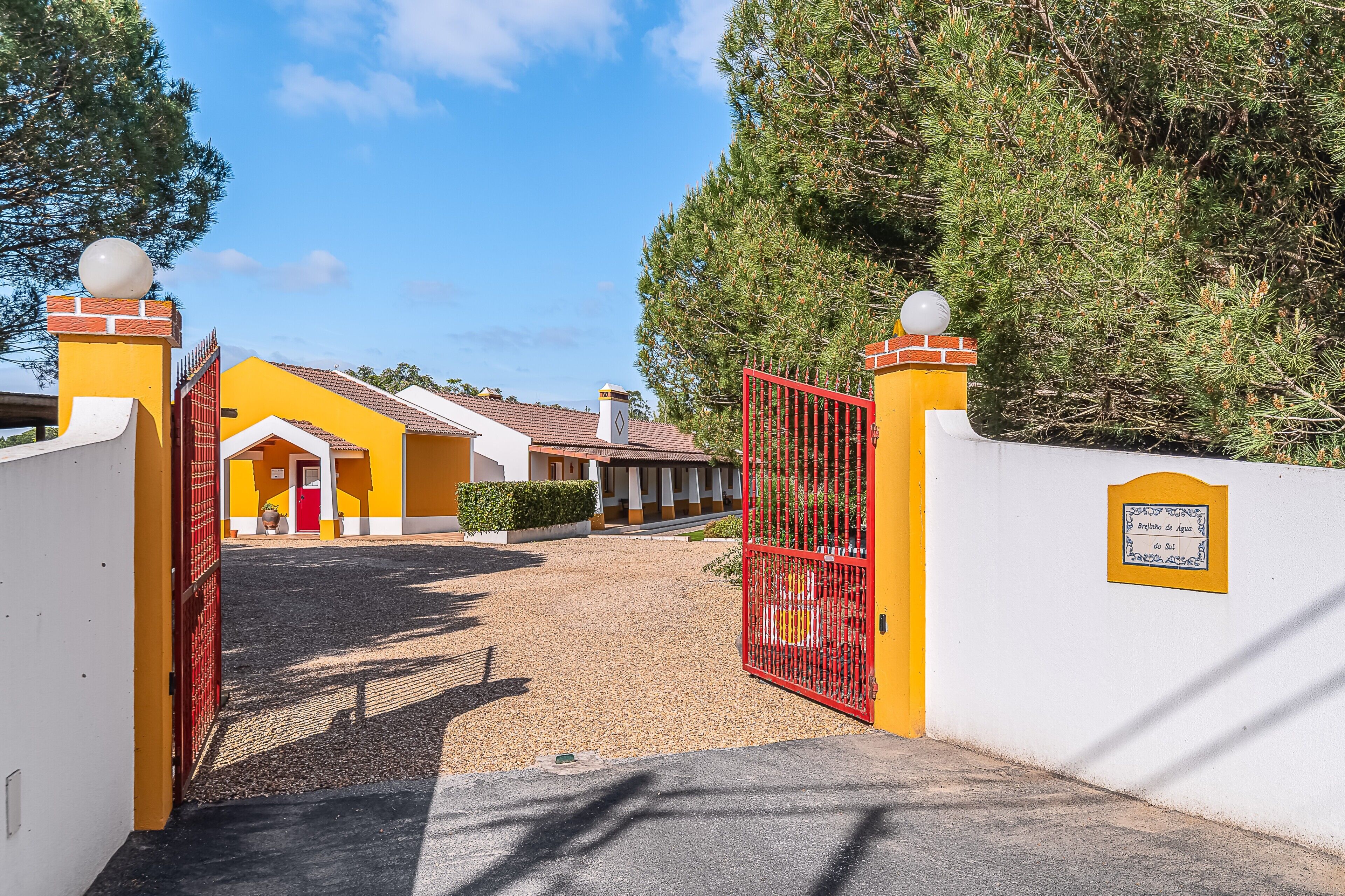 A gated entrance to a building with a yellow facade, a red gate, and a gravel driveway leading to the entrance.