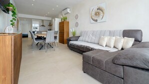 Una sala moderna con un sofá gris, un comedor con sillas, una cocina al fondo y un aire acondicionado instalado en la pared.