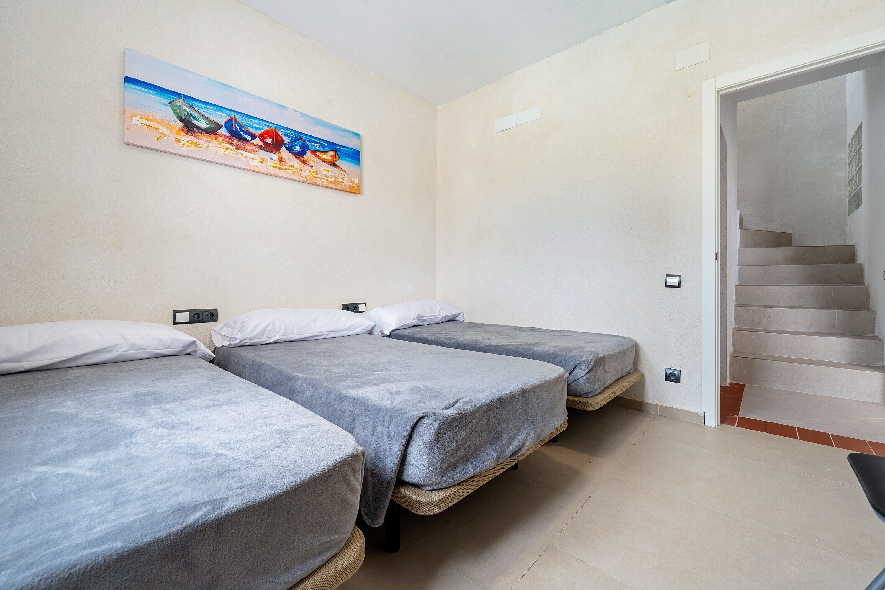 A room with three single beds, a painting of boats on the wall, and a staircase leading to an upper floor.