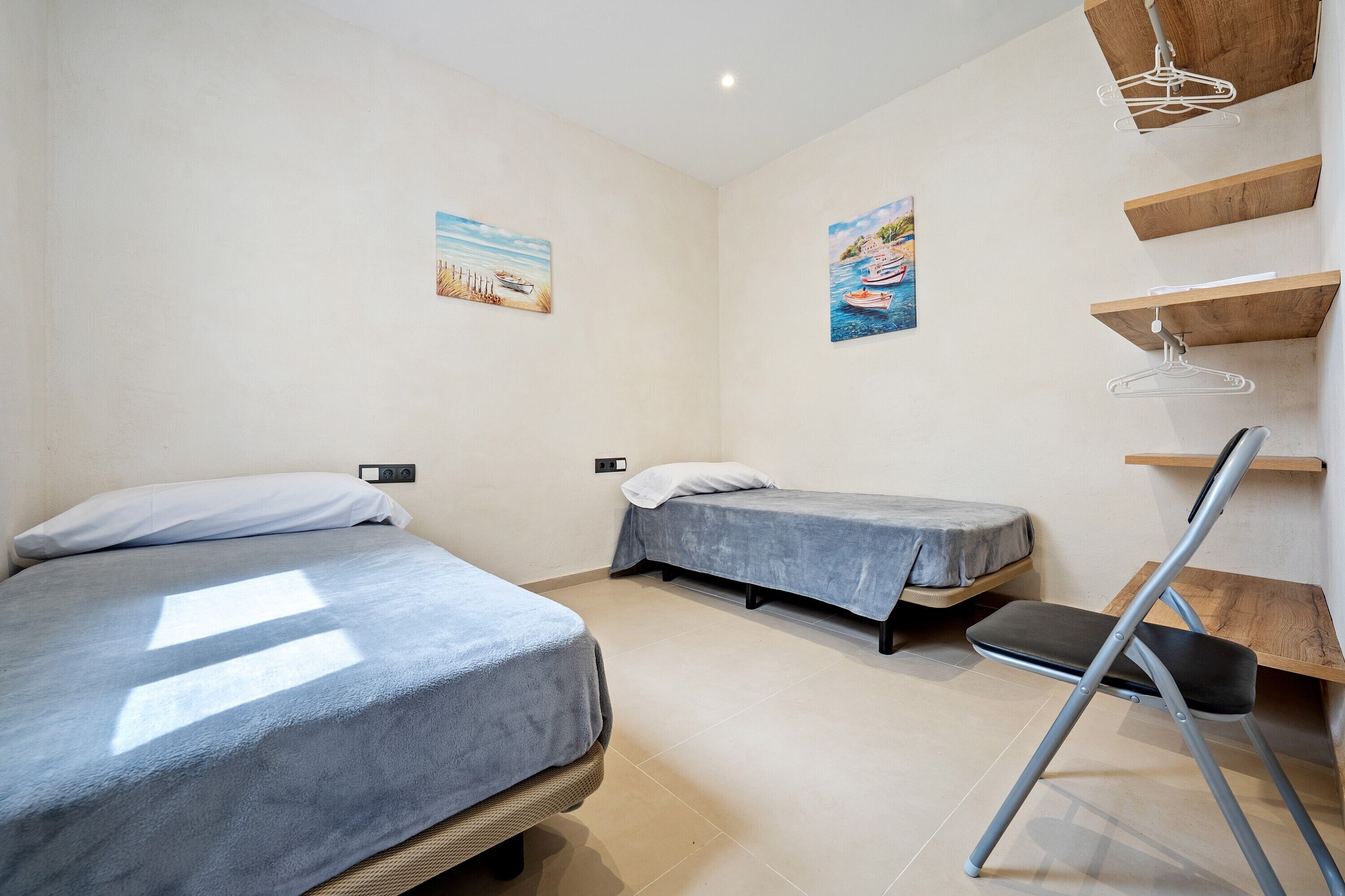 A room with two beds, a desk, and a chair. There are wall-mounted shelves and a hanging coat rack.