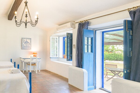 A room with two beds, a desk with a chair, a chandelier, and a large window with curtains.