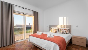 Une chambre à coucher comprenant un lit, une table de chevet, une porte-fenêtre coulissante donnant sur une terrasse et une vue sur un immeuble.