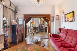 A living room with a red sofa, a wooden coffee table, a vintage clock, and a chandelier. - Room 'Quarto Duplo Com Varanda' with Balcony, Wi-Fi and Air Conditioning (São Brás de Alportel)