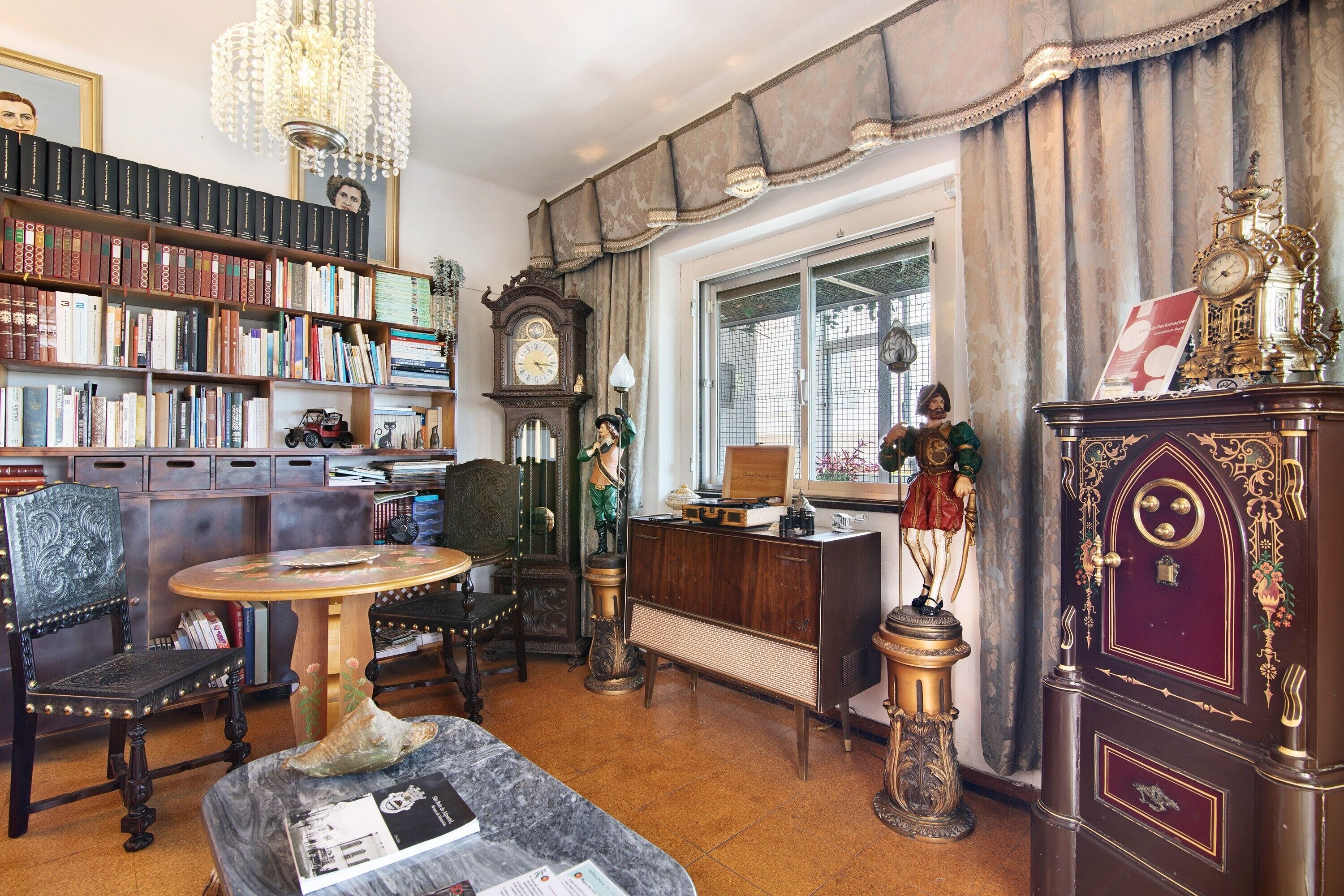 A room with a large bookshelf, a wooden cabinet with decorative items, a round table, and a vintage clock.