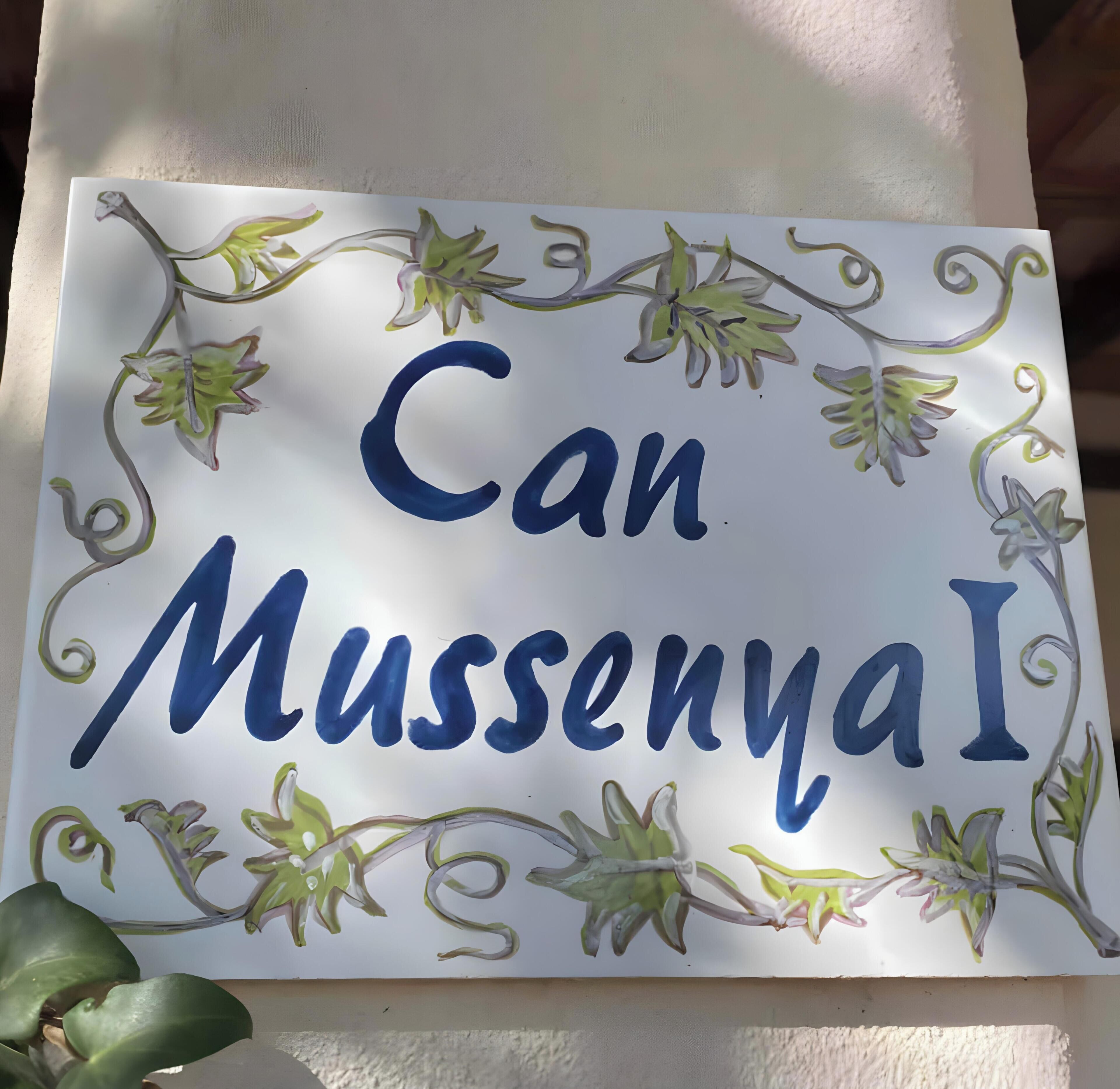 Ein Schild mit dem Text 'Can MussengaI', umgeben von dekorativen floralen Elementen.