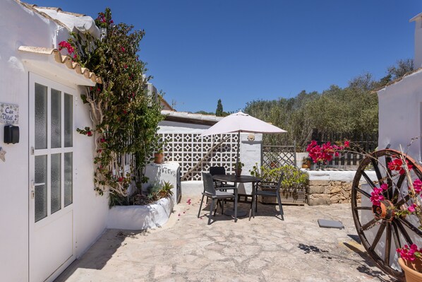 A courtyard with a white building, a table with chairs, a wheelbarrow, and potted plants.