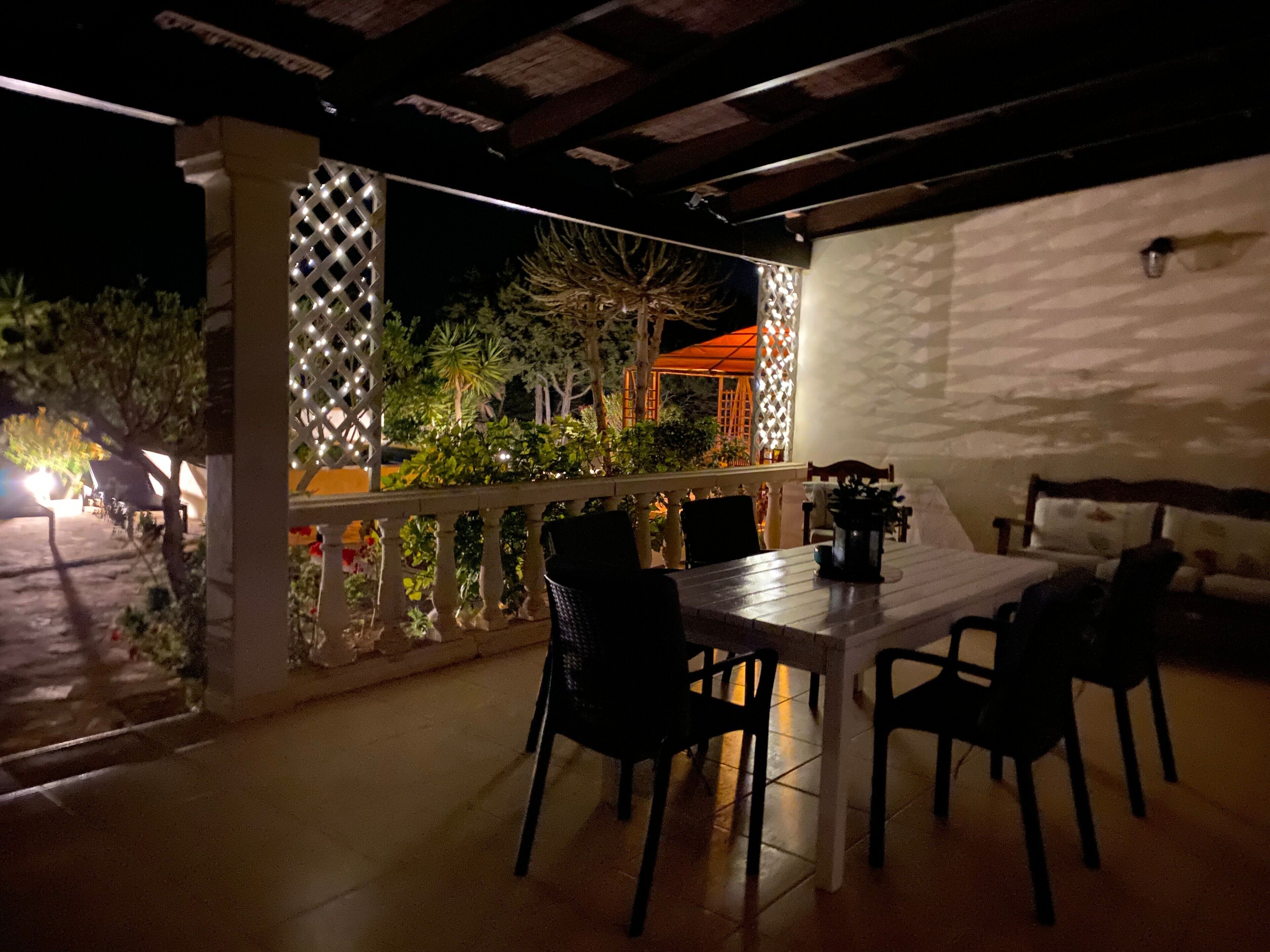 Ein Terrassenbereich im Freien mit Tisch und Stühlen, einer weißen Wand und einem Gitterzaun.