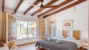 Une chambre à coucher comprenant un lit, un ventilateur de plafond, une fenêtre avec des rideaux, une table de chevet et une lampe fixée au mur.