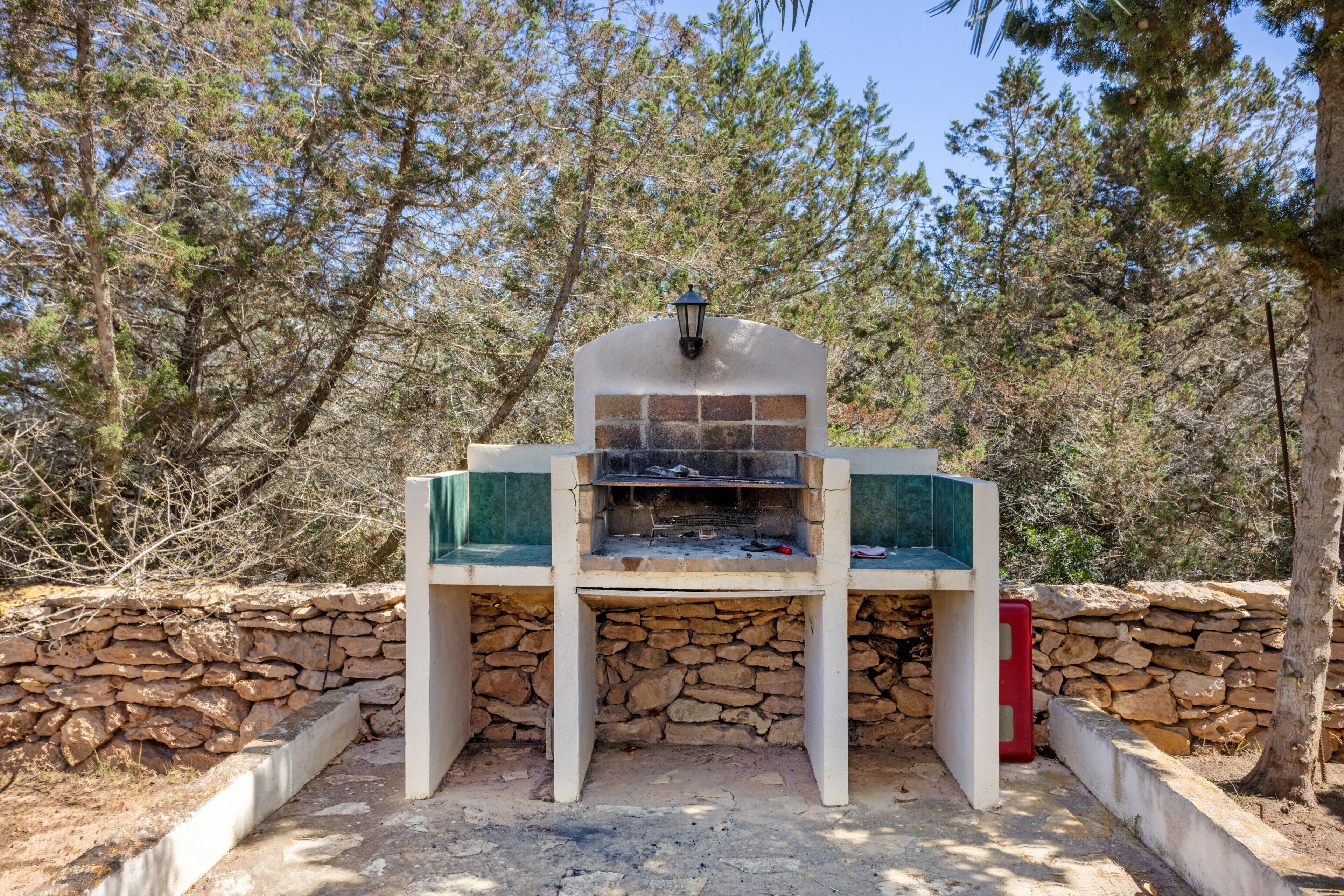 Una parrilla de piedra al aire libre con un farol encima, rodeada por un muro de piedra y árboles.